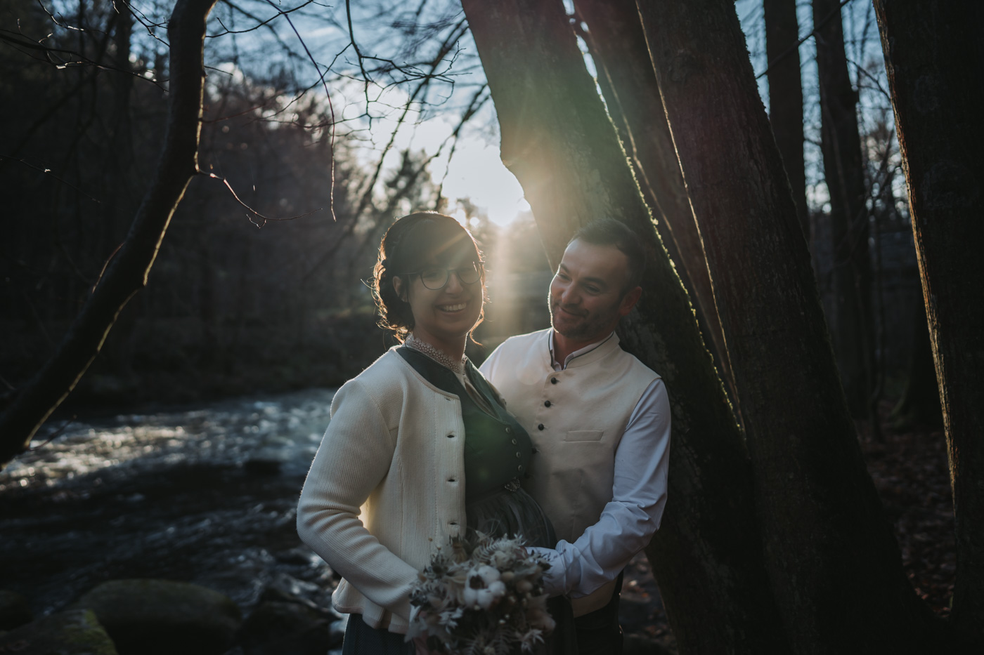 Brautpaar steht Arm in Arm, von vorne betrachtet, vor einem ruhigen Fluss in der Abendsonne. Ihre Gesichter strahlen Glück und Liebe aus, während die untergehende Sonne ein warmes, goldenes Licht auf sie wirft.